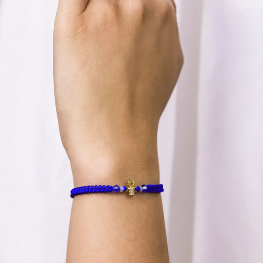 Mano de mujer con pulsera tejida en hilo azul, accesorios de piedras de color azul y blanco, y dije en forma de niño en oro amarillo de 18 quilates con cristales de color blanco, hecho en Joyería Caracas.