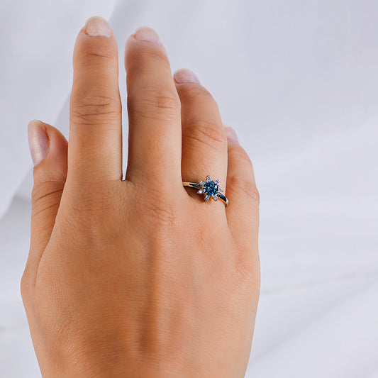 Mano de mujer con anillo en forma de flor en oro blanco de 18 quilates con cristal de color azul en el centro y blancos alrededor, hecho en Joyería Caracas.