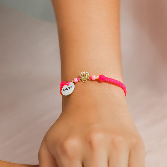 Mano de mujer con pulsera tejida en hilo fucsia coreano, bisutería y dije con forma de corona en oro amarillo de 18 quilates con cristales de color blanco, hecho en Joyería Caracas.