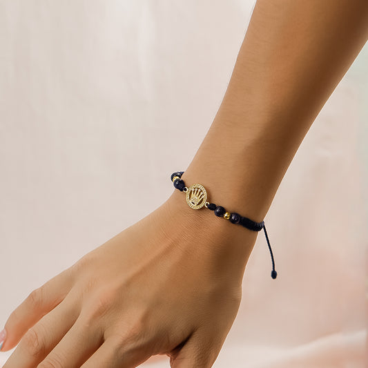 Mano de mujer con pulsera tejida en hilo negro, bisutería y dije con forma de corona en oro amarillo de 18 quilates con cristales de color blanco, hecho en Joyería Caracas.