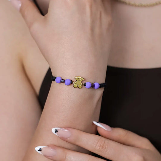 Mano de mujer con pulsera tejida en hilo negro con accesorios en color morado y dije de osito en oro amarillo de 18 quilates, hecho en Joyería Caracas.