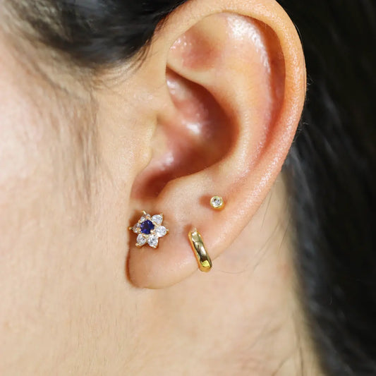 Oreja de mujer usando unos topitos en forma de flor en oro amarillo de 18 quilates con un cristal de color azul en el centro y cristales de color blanco alrededor de Joyería Caracas.
