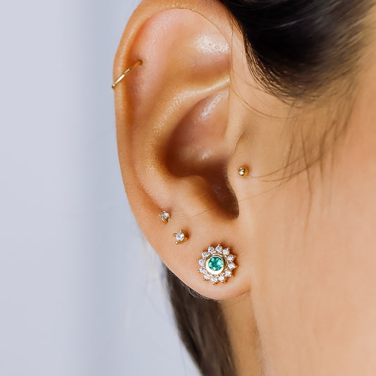 Oreja de mujer usando topito en forma de flor en oro amarillo de 18 quilates con un cristal de color verde en el centro y cristales de color blanco alrededor.