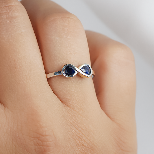 Mano de mujer usando anillo con forma de infinito en el centro en oro blanco de 18 quilates y 2 zafiros de color azul en forma de gota, hecho en Joyería Caracas.