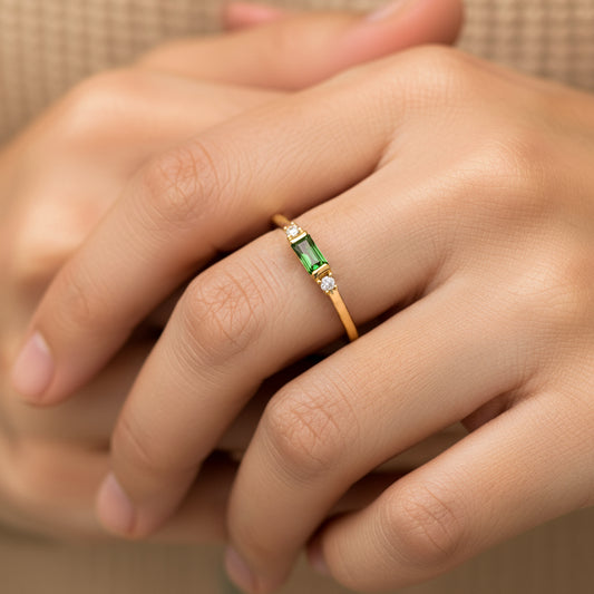 Mano de mujer usando un anillo en oro amarillo de 18 quilates con cristal rectangular de color verde en el centro con 2 cristales a cada lado de color blanco de Joyería Caracas.