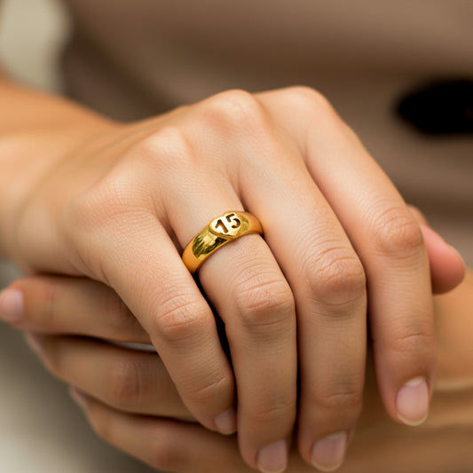 Mano de mujer usando un anillo plano con el número 15 dentro de un corazón en el centro en oro amarillo de 18 quilates de Joyería Caracas.
