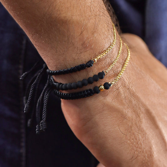 Mano de hombre con pulsera tejida en hilo color negro y cadena en tejido bismarck en oro amarillo de 18 quilates, hecho en Joyería Caracas.