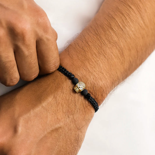 Mano de hombre con pulsera tejida en hilo negro terlenca, accesorios en bisutería de color negro y dije en forma de calavera en oro amarillo de 18 quilates con cristales de color blanco, hecho en Joyería Caracas.