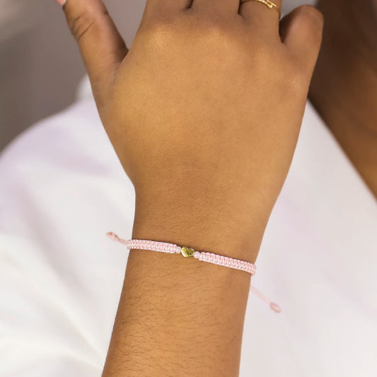 Mano de mujer con pulsera tejida en hilo rosa claro, accesorios de bisutería de color rosa y dije en forma de corazón en oro amarillo de 18 quilates, hecho en Joyería Caracas.