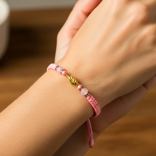Mano de modelo usando una pulsera tejida en hilo color rosa, herraje en forma de ala en oro amarillo de 18 quilates y bisutería de calidad de Joyería Caracas.
