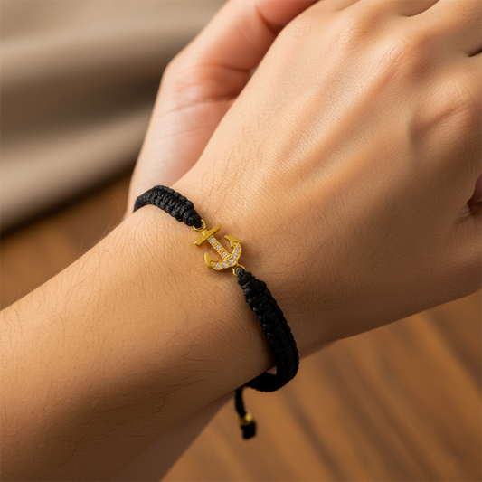Mano de mujer usando una pulsera tejida en hilo de color negro con herraje en forma de ancla en oro amarillo de 18 quilates con cristales de color blanco de Joyería Caracas.