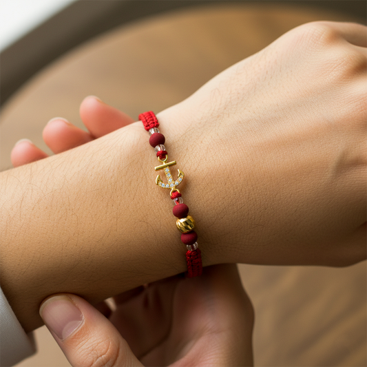 Mano de modelo usando una pulsera tejida de hilo color rojo, herraje en forma de ancla en oro amarillo de 18 quilates con cristales de color blanco, esfera facetada y bisutería de calidad de Joyería Caracas.