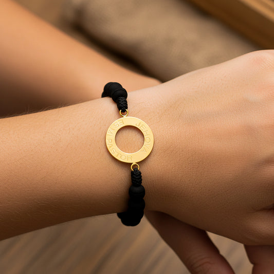 Mano de modelo usando una pulsera tejida en hilo color negro con herraje circular con la palabra Jehova es mi pastor en oro amarillo de 18 quilates de Joyería Caracas.