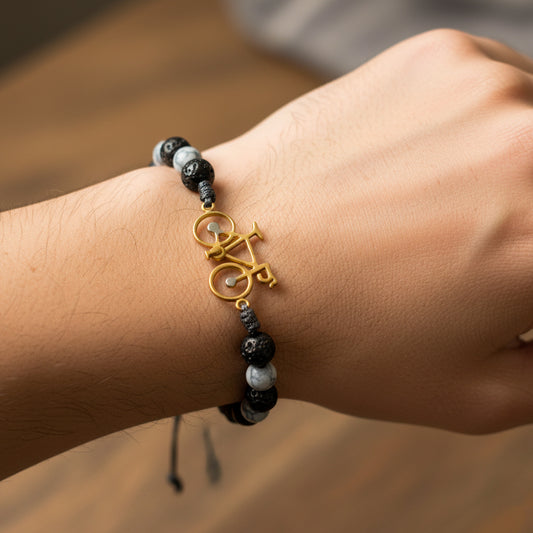 Mano de modelo usando una pulsera tejida en hilo de color gris con dije en forma de bicicleta en oro amarillo de 18 quilates con bisutería de calidad de Joyería Caracas.