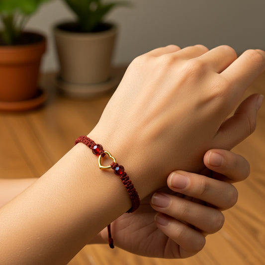 Mano de modelo usando una pulsera tejida en hilo color vinotinto con dije en forma de corazón en oro amarillo de 18 quilates y bisutería de calidad de Joyería Caracas.