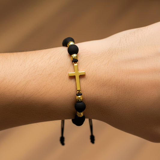 Mano de modelo usando una pulsera tejida en hilo de color negro con dije de cruz con esferas facetadas en oro amarillo de 18 quilates con bisutería de calidad de Joyería Caracas.