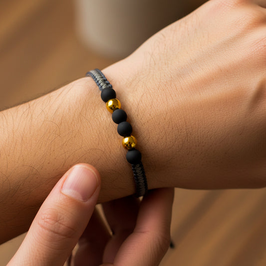 Mano de hombre usando una pulsera tejida en hilo combinado de color gris claro y oscuro con bisutería de color negro con 2 esferas lisas en oro amarillo de 18 quilates de Joyería Caracas.