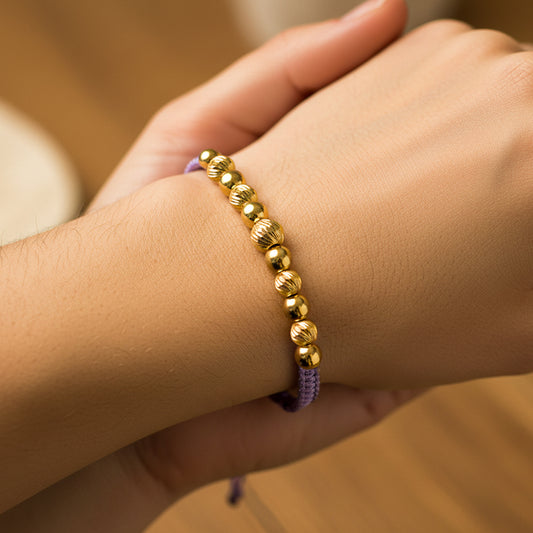 Mano de mujer usando una pulsera tejida de hilo color lila con esferas facetadas y lisas en oro amarillo de 18 quilates de Joyería Caracas.