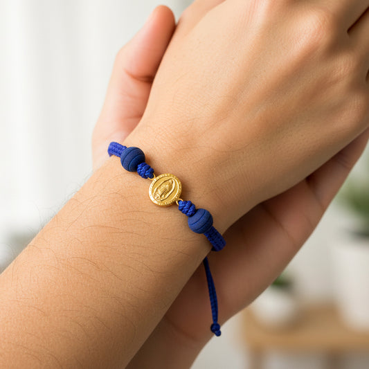 Mano de modelo usando una pulsera tejida en hilo color azul oscuro con dije de la Virgen de Guadalupe en oro amarillo de 18 quilates y bisutería de calidad de Joyería Caracas.