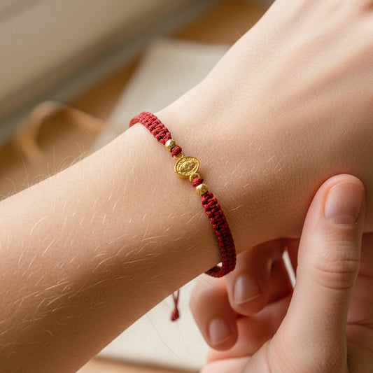 Mano de modelo usando una pulsera tejida en hilo color rojo con herraje de la Virgen Milagrosa con esferas lisas en oro 18 quilates de Joyería Caracas.