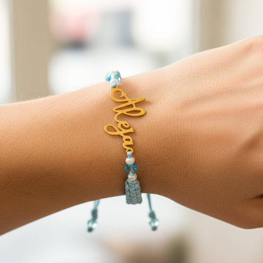 Mano de modelo usando una pulsera tejida en hilo color celeste con dije de nombre personalizado en oro amarillo de 18 quilates de Joyería Caracas.