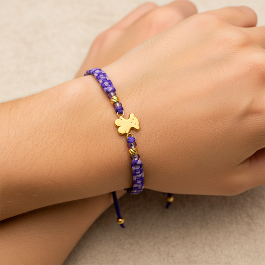 Mano de modelo usando una pulsera tejida en hilo color morado con herraje en forma de osito y 2 esferas facetadas en oro amarillo de 18 quilates con bisutería de calidad de Joyería Caracas.