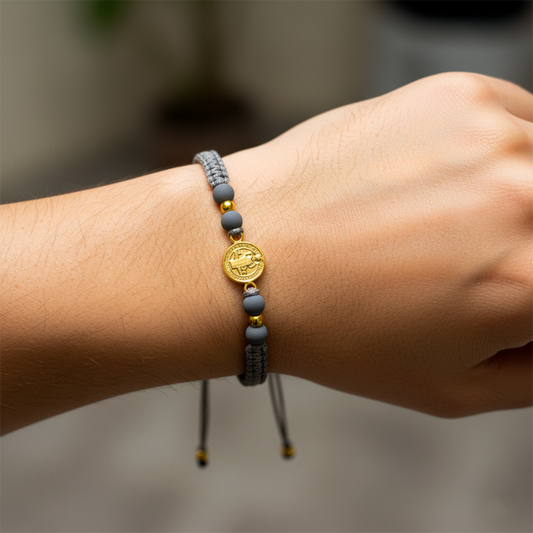Mano de modelo usando una pulsera tejida en hilo color gris con dije de San Benito y 2 esferas lisas en oro amarillo de 18 quilates con bisutería de calidad de Joyería Caracas.
