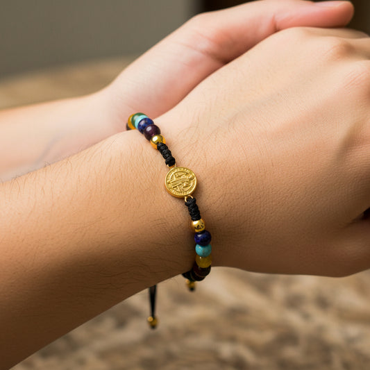 Mano de modelo usando una pulsera tejida en hilo color negro con dije de San Benito y 2 esferas lisas en oro amarillo de 18 quilates y bisutería de calidad de Joyería Caracas.