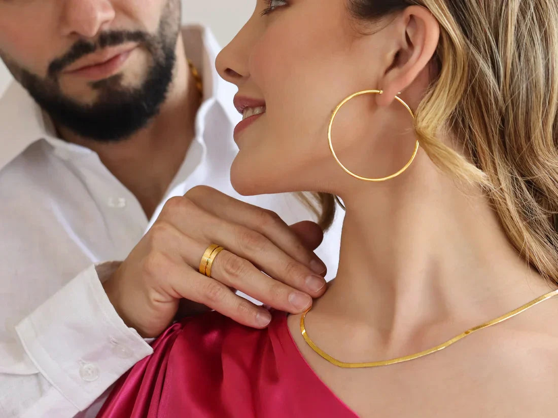 Un hombre usa en su dedo anular un anillo y acomoda una gargantilla tipo espejo en el cuello de una mujer con una candonga en oro