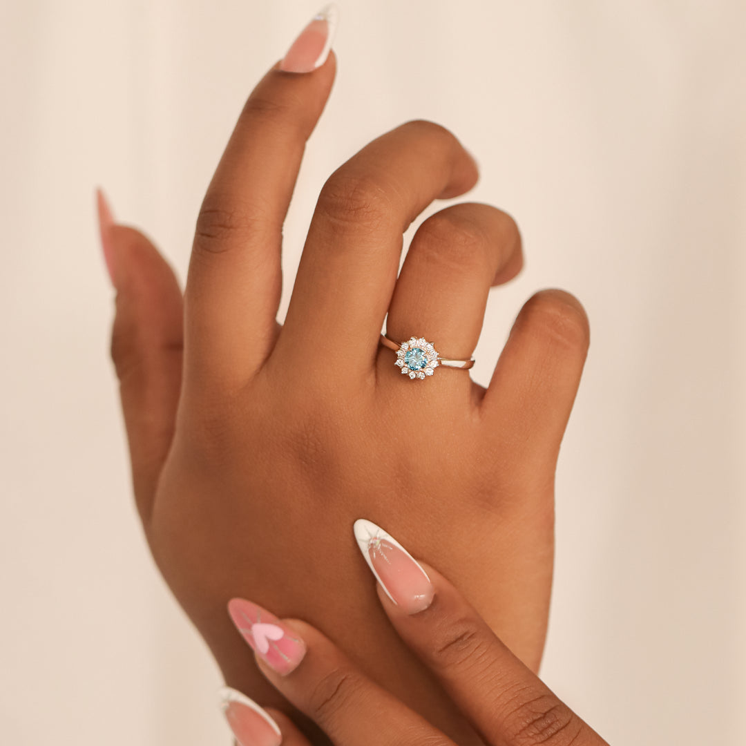 Mano de mujer con anillo inspirado en las palmeras de Miami, en oro blanco de 18 quilates con cristales de color blanco alrededor y celeste en el centro.