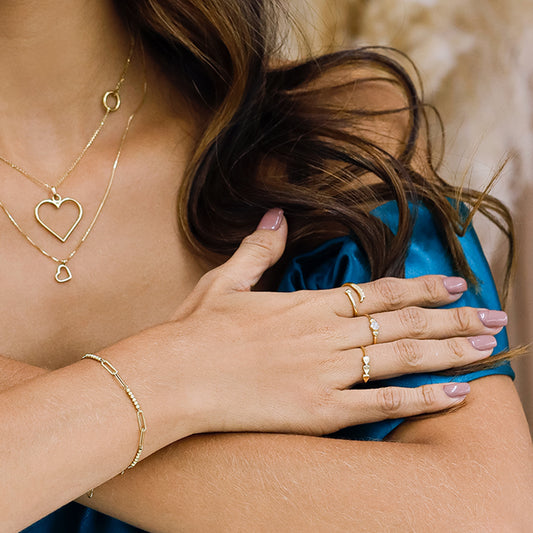 Mano de mujer usando anillo de circulo en el centro con cristales de color blanco y 1 corazón en cada lado en oro amarillo de 18 quilates.
