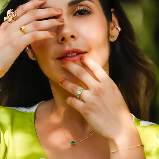 Mano de mujer usando un anillo en oro amarillo de 18 quilates con Diamante en el centro en forma redonda de color blanco y 2 Esmeraldas en forma de gota de color verde de Joyería Caracas. 