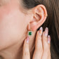 Oreja de mujer usando un arete en forma cuadrada en oro amarillo de 18 quilates con un cristal de color verde en el centro en forma cuadrada y cristales de color blanco alrededor de Joyería Caracas. 