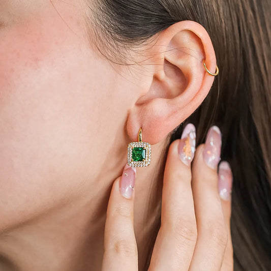 Oreja de mujer usando un arete en forma cuadrada en oro amarillo de 18 quilates con un cristal de color verde en el centro en forma cuadrada y cristales de color blanco alrededor de Joyería Caracas. 