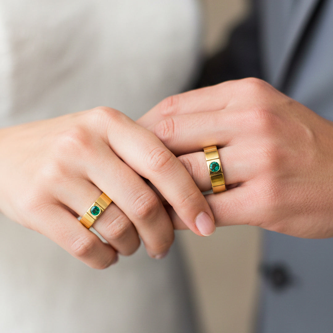 Manos de una pareja usando unas argollas de matrimonio en oro amarillo de 18 quilates con cristal verde de Joyería Caracas. 