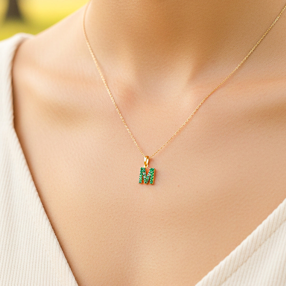 Cuello de modelo usando una cadena con un dije en forma de la letra M en oro amarillo de 18 quilates con cristales verdes de Joyería Caracas.