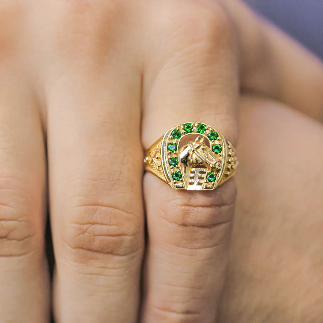 Mano de hombre con anillo en forma de herradura con caballo en el centro en oro amarillo de 18 quilates y cristales de color verde alrededor, hecho en Joyería Caracas. 