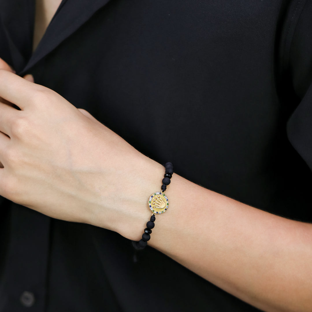Mano de hombre con pulsera tejida en hilo negro con dije circular con corona en el centro en oro amarillo de 18 quilates con cristales de color blanco, verde y azul, hecho en Joyería Caracas. 