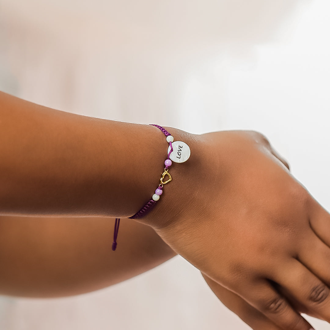 Mano de mujer con pulsera tejida en hilo morado, bisutería y dije en forma de corazón en oro amarillo de 18 quilates, hecho en Joyería Caracas. 