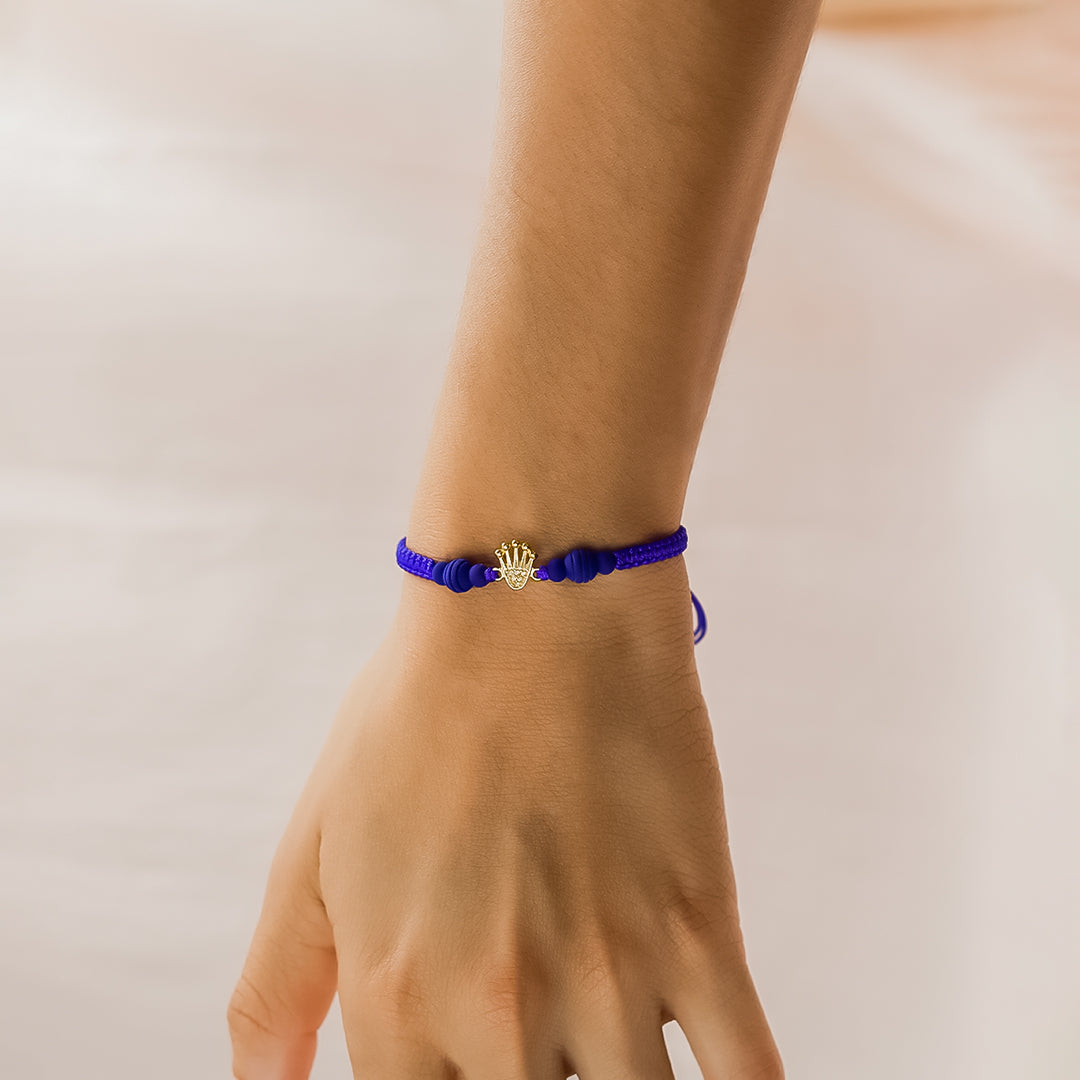 Mano de mujer con pulsera tejida en hilo azul coreano, bisutería y dije con forma de corona en oro amarillo de 18 quilates con cristales de color blanco, hecho en Joyería Caracas. 
