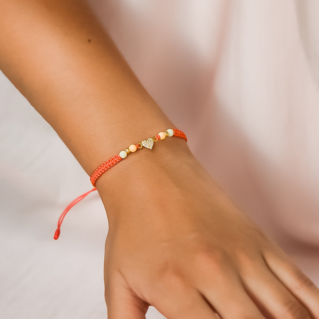 Mano de mujer con pulsera tejida en hilo color coral, bisutería y dije en forma de corazón en oro amarillo de 18 quilates con cristales de color blanco, hecho en Joyería Caracas. 