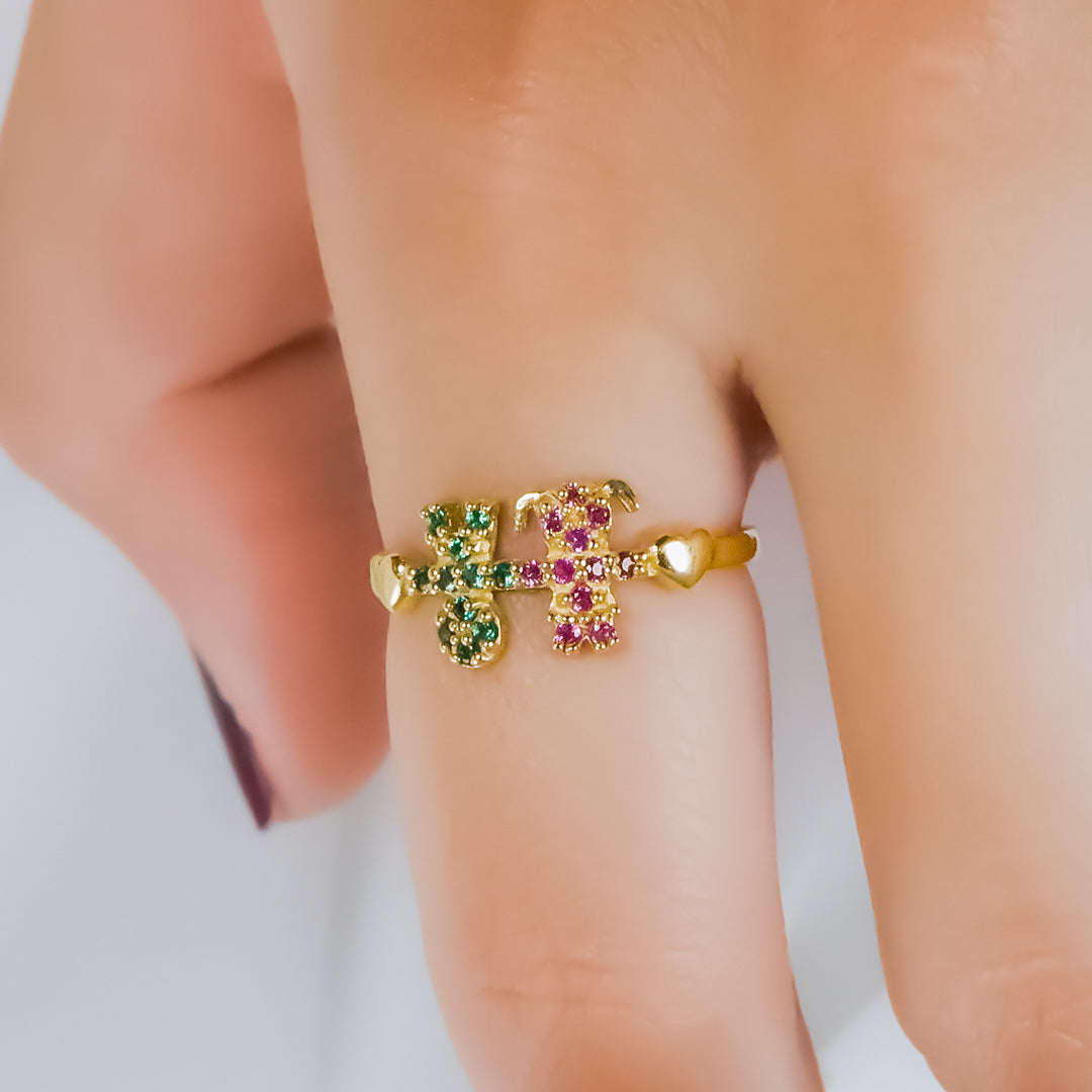 Mano de mujer usando anillo con forma de niño y niña en oro amarillo de 18 quilates con cristales de color verde y rosa, hecho en Joyería Caracas. 