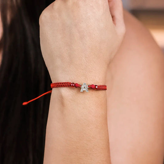 Mano de mujer con pulsera tejida en hilo rojo, accesorios en color rojo y dije de la letra A en oro amarillo de 18 quilates, hecho en Joyería Caracas. 