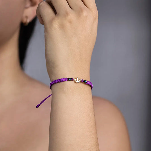 Mano de mujer con pulsera tejida en hilo morado, accesorios en color morado, azul celeste y dije de corona en oro amarillo de 18 quilates, hecho en Joyería Caracas. 