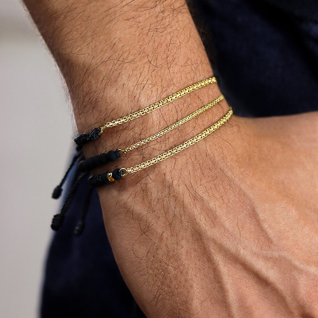 Mano de hombre con pulsera tejida en hilo color negro y cadena en tejido bismarck en oro amarillo de 18 quilates, hecho en Joyería Caracas. 