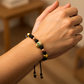Mano de mujer usando una pulsera tejido en hilo de color negro con bisutería del mismo color, con esferas 3 oros facetadas y dije con forma de calavera en oro amarillo de 18 quilates con 2 Esmeraldas de color verde de Joyería Caracas. 