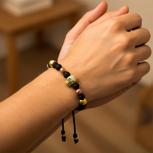 Mano de mujer usando una pulsera tejido en hilo de color negro con bisutería del mismo color, con esferas 3 oros facetadas y dije con forma de calavera en oro amarillo de 18 quilates con 2 Esmeraldas de color verde de Joyería Caracas. 