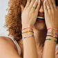 Mano de mujer usando una pulsera tejida en hilo azul celeste y accesorios del mismo color y herraje en el centro de la palabra Dios en oro amarillo de 18 quilates con cristales de color blanco en él de Joyería Caracas.