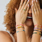 Mano de mujer usando una pulsera tejida en hilo de color gris con accesorios de color rojo y amarillo y esfera lisa en el centro en oro amarillo de 18 quilates de Joyería Caracas. 