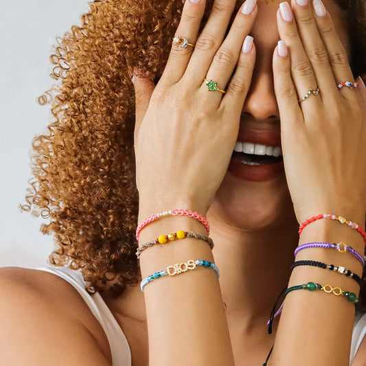 Mano de mujer usando una pulsera tejida en hilo de color gris con accesorios de color rojo y amarillo y esfera lisa en el centro en oro amarillo de 18 quilates de Joyería Caracas. 